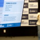 Foto mostra televisão em primeiro plano com informações sobre o programa saúde conectada. Ao fundo, há uma mulher no palco, falando em um microfone