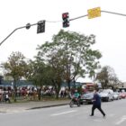 Foto mostra homem atravessando uma rua pavimentada na faixa de pedestres, enquanto uma moto e carros aguardam. No alto, há um semáforo no estágio vermelho para os veículos
