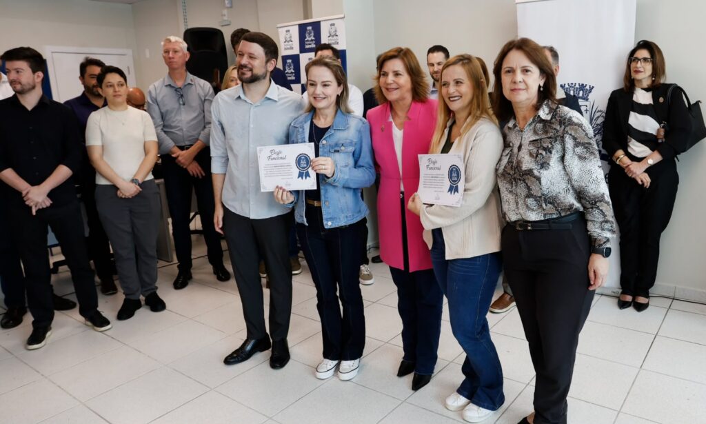 Foto mostra, em primeiro plano, quatro mulheres e um homem em pé, sorrindo para a foto. Duas delas exibe um documento. Atrás do grupo, há outras pessoas em pé