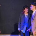 Foto mostra dois homens e uma mulher jovens usando roupas antigas em uma cena de teatro